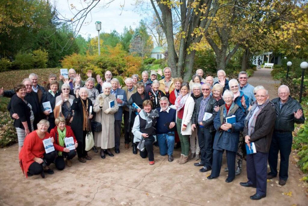 A large group of people gather for a group shot outside amongst autumn trees and a garden pathway.