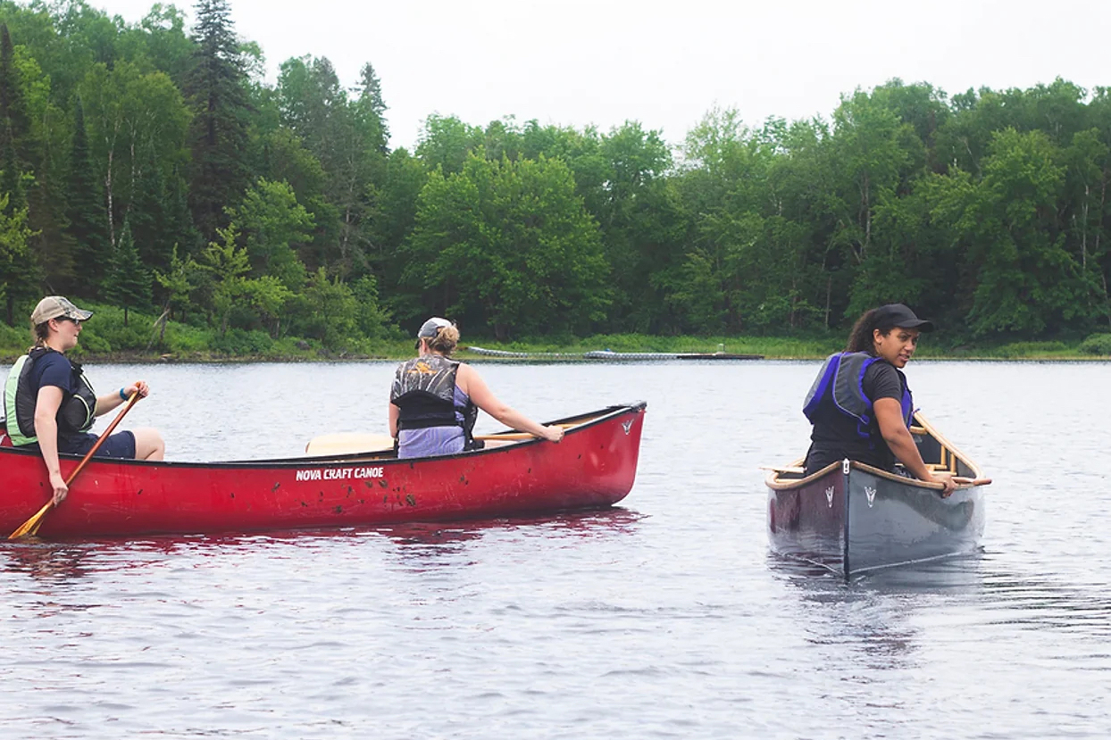 Two women paddling in a red canoe. Beside them, a woman in a dark blue canoe looks back at the camera.