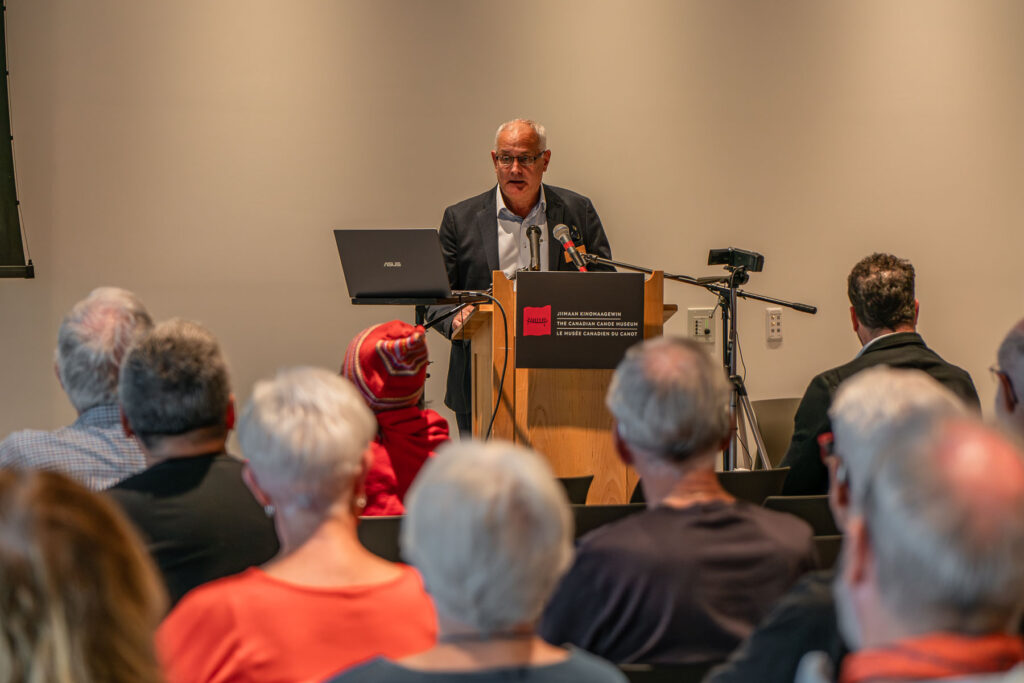 Kevin Malone, chair of the Canadian Canoe Museum, stands at a podium addressing a room of people during the 2024 Annual General Meeting. The backs of heads are shown.