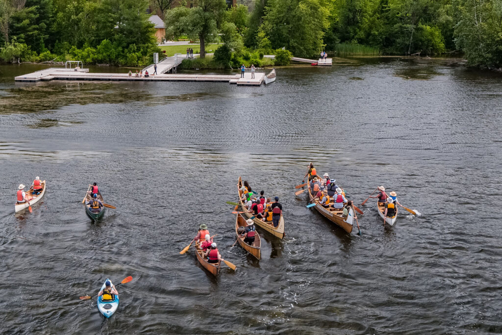 An overhead shot of a flotilla of canoes, including a voyageur canoe, approach the docks of The Canadian Canoe Museum on Little Lake during National Canoe Day (June 26, 2024).