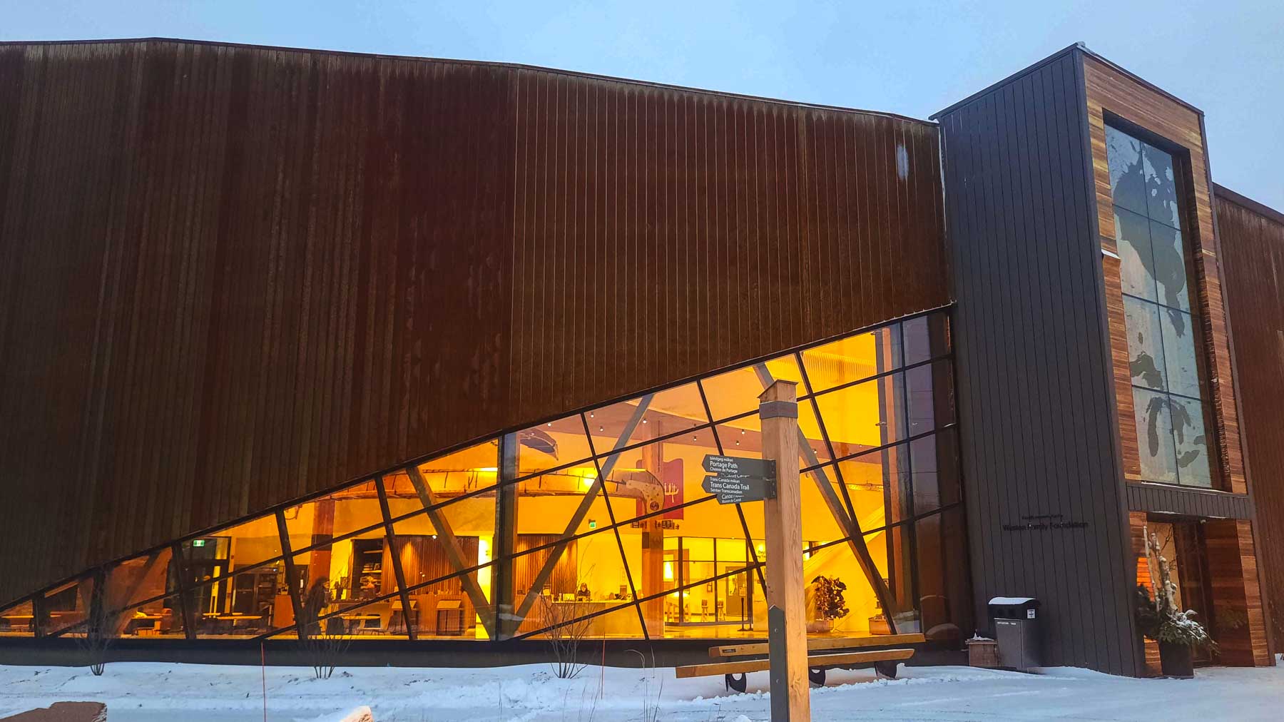 The exterior front entrance of the Canadian Canoe Museum. Snow lays on the ground and warm light floods out from the glass windows.