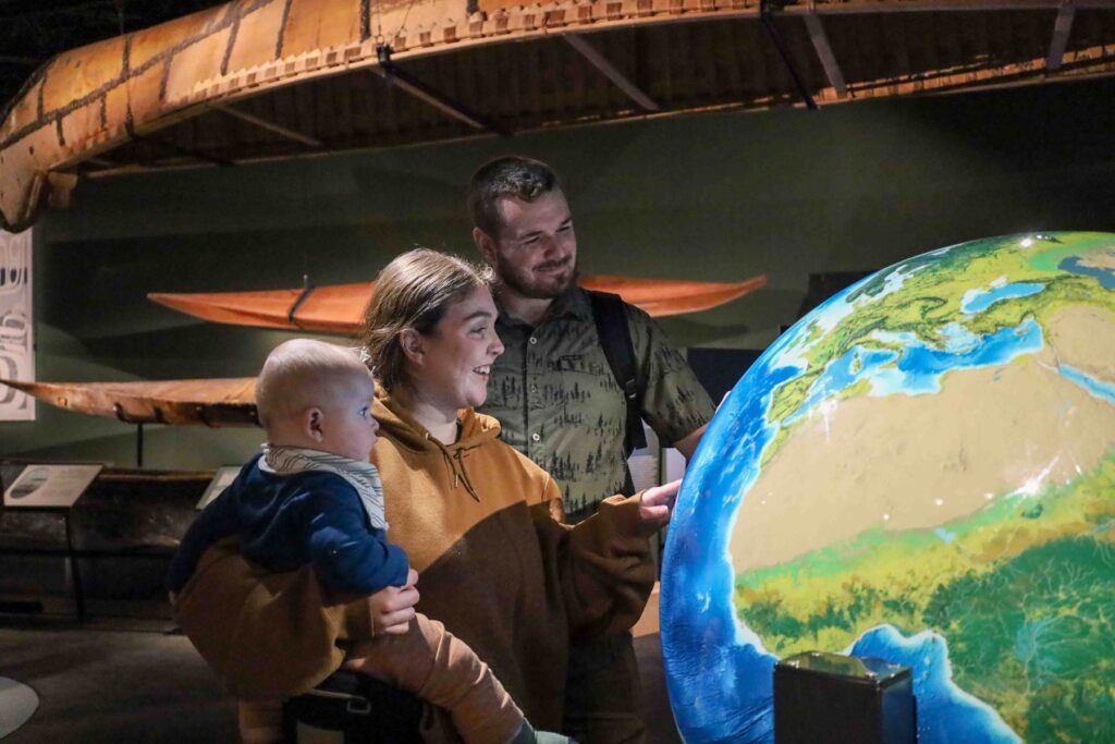 A family holding a baby stands in the Exhibition Hall in front of a large globe, looking on in amazement. Canoes hang in the background.