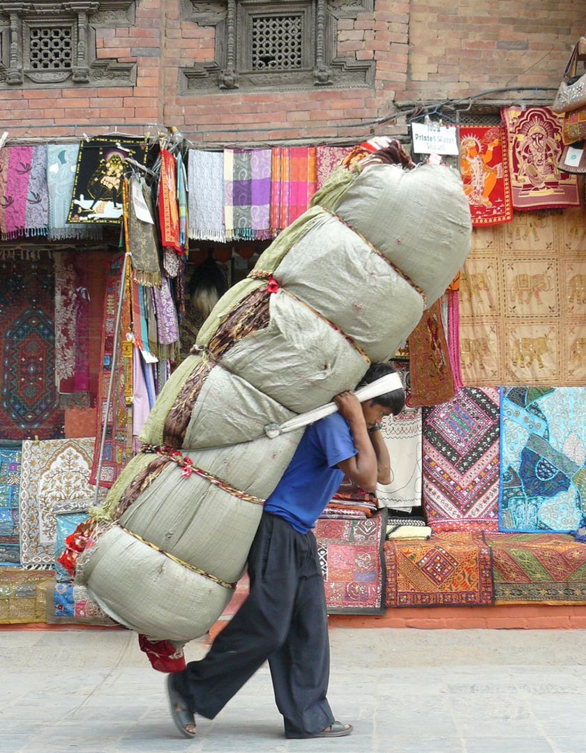 A Nepalese porters carries a large load on his back while walking down the street in front of a colourful display of rugs.