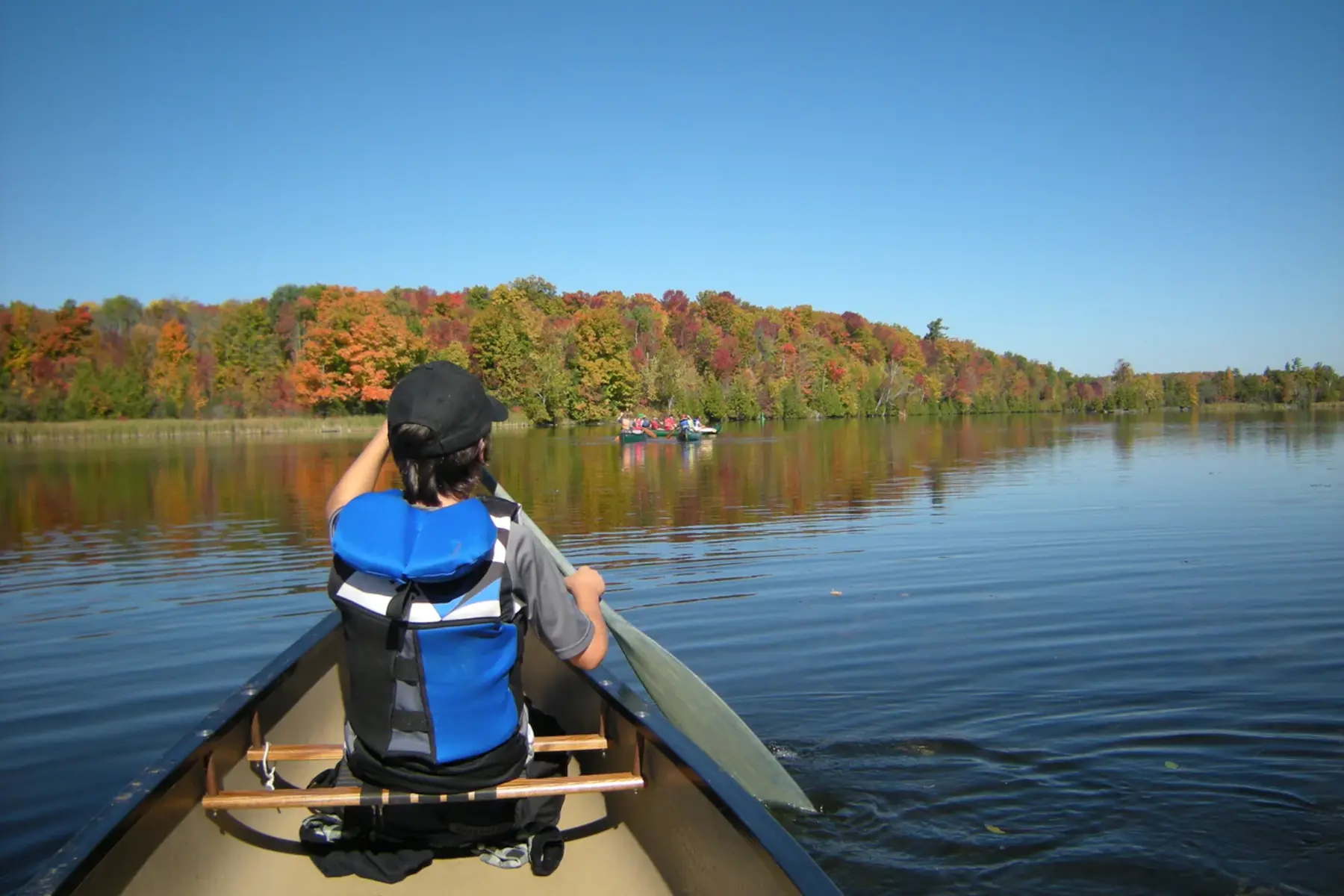 Paddler at the front of the canoe facing away from the camera paddles through a calm lake on a sunny fall day.