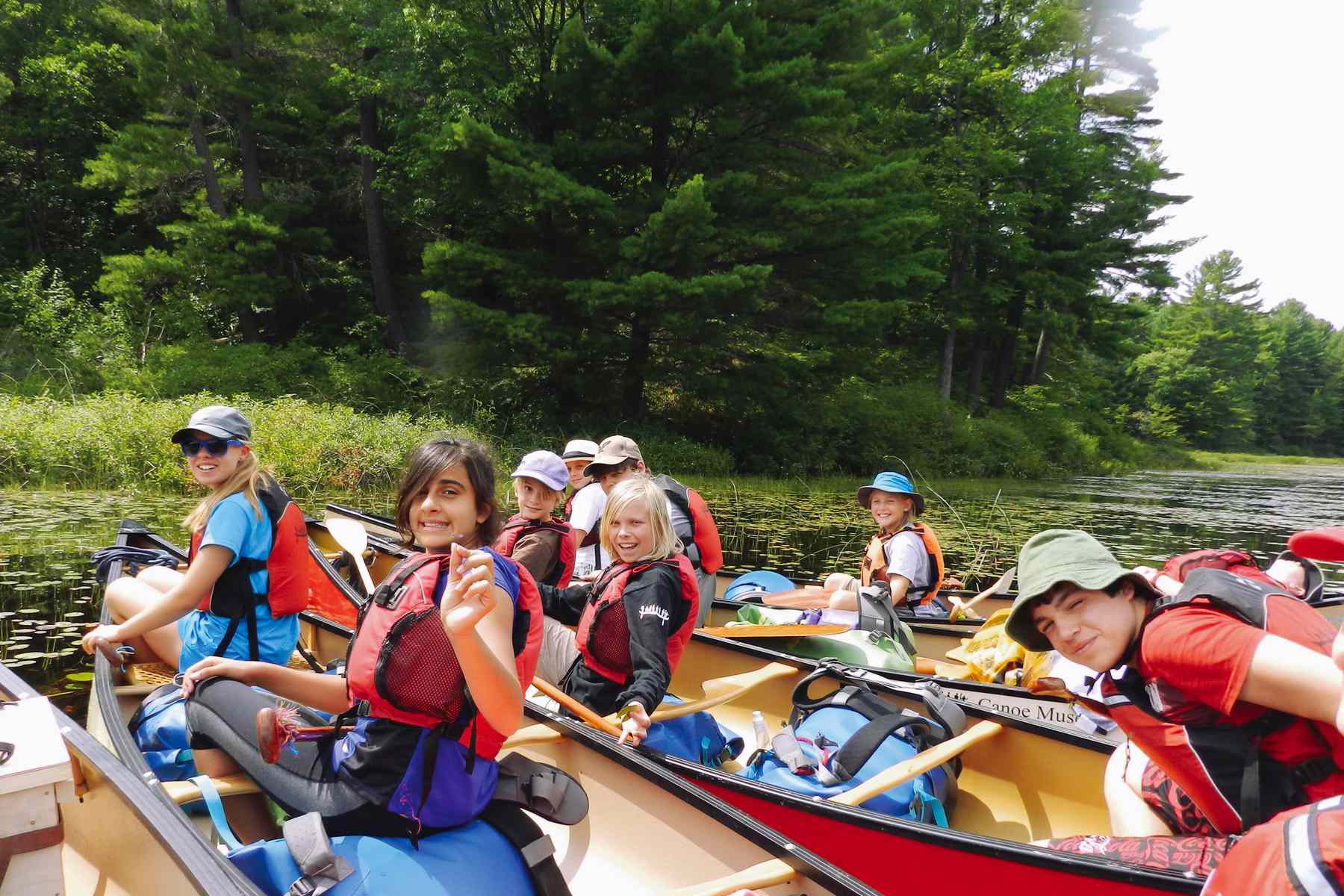 A group of youth and their instructors in three canoes look back and smile and wave at the camera. Their canoes are filled with packs for a canoe trip.
