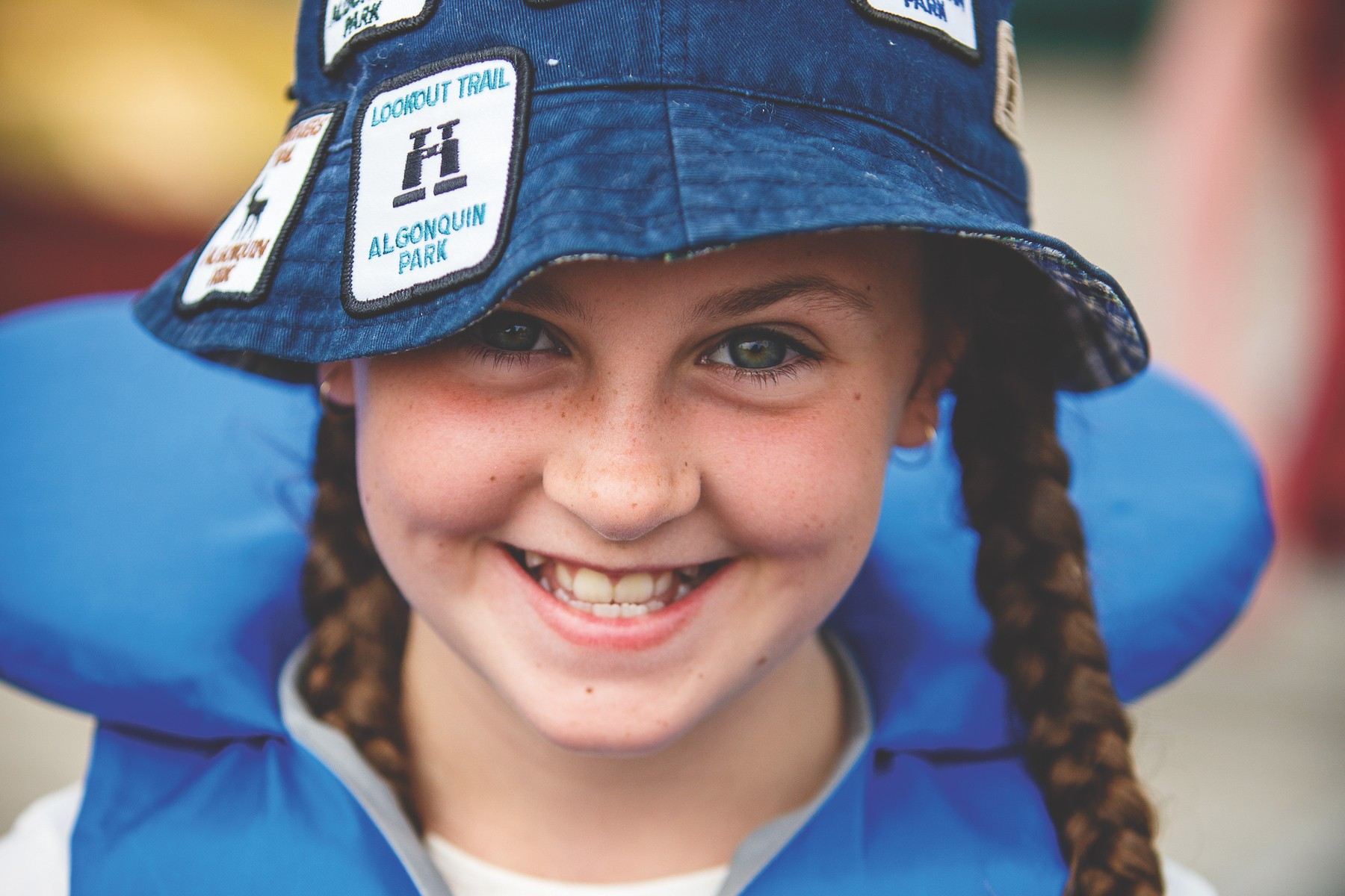A young girl with braids is wearing a blue personal flotation device (PFD) and a blue bucket hat that is decorated with various sew-on patches depicting different trails in Algonquin Park. She is smiling and looking straight at the camera.