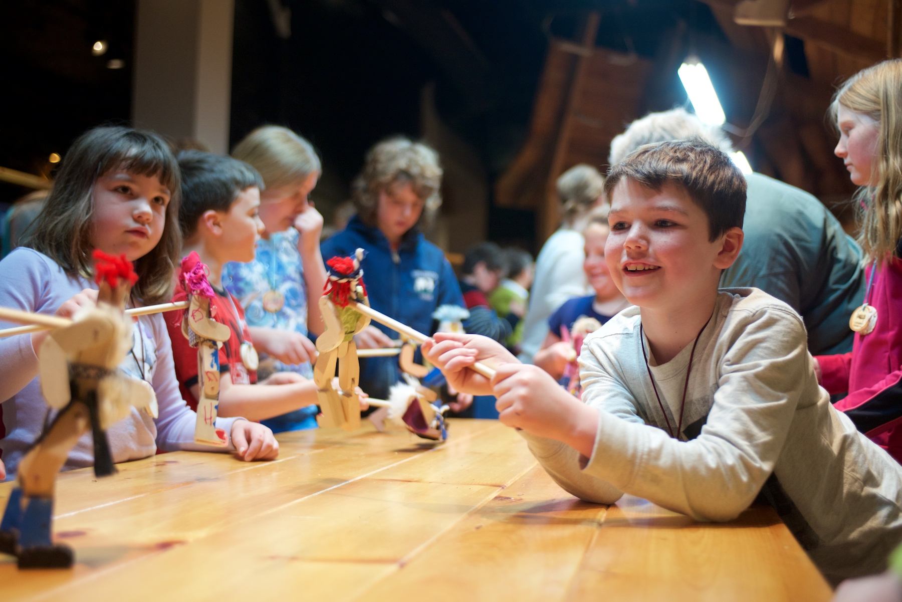 A young boy smiles while making his wooden limberjack toy that he decorated dance. He is surrounded by other children playing with their own limberjack toys.