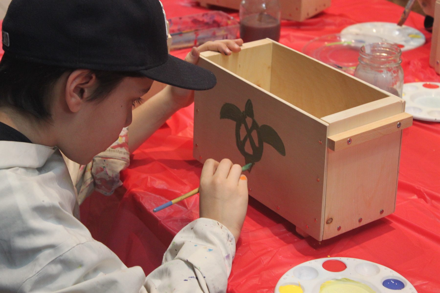 A young boy concentrates as he paints a green turtle onto a wooden wanigan or box. There is a paint try beside him.