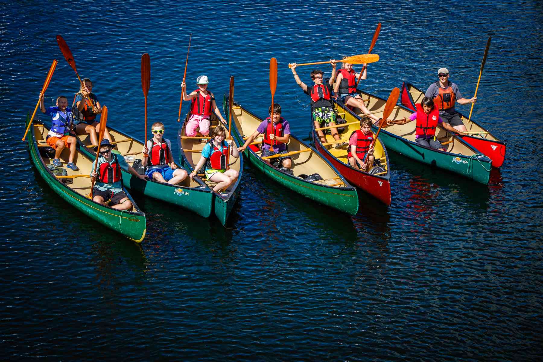 Campers and their instructors all raise their paddles to the camera in seven side by side canoes.