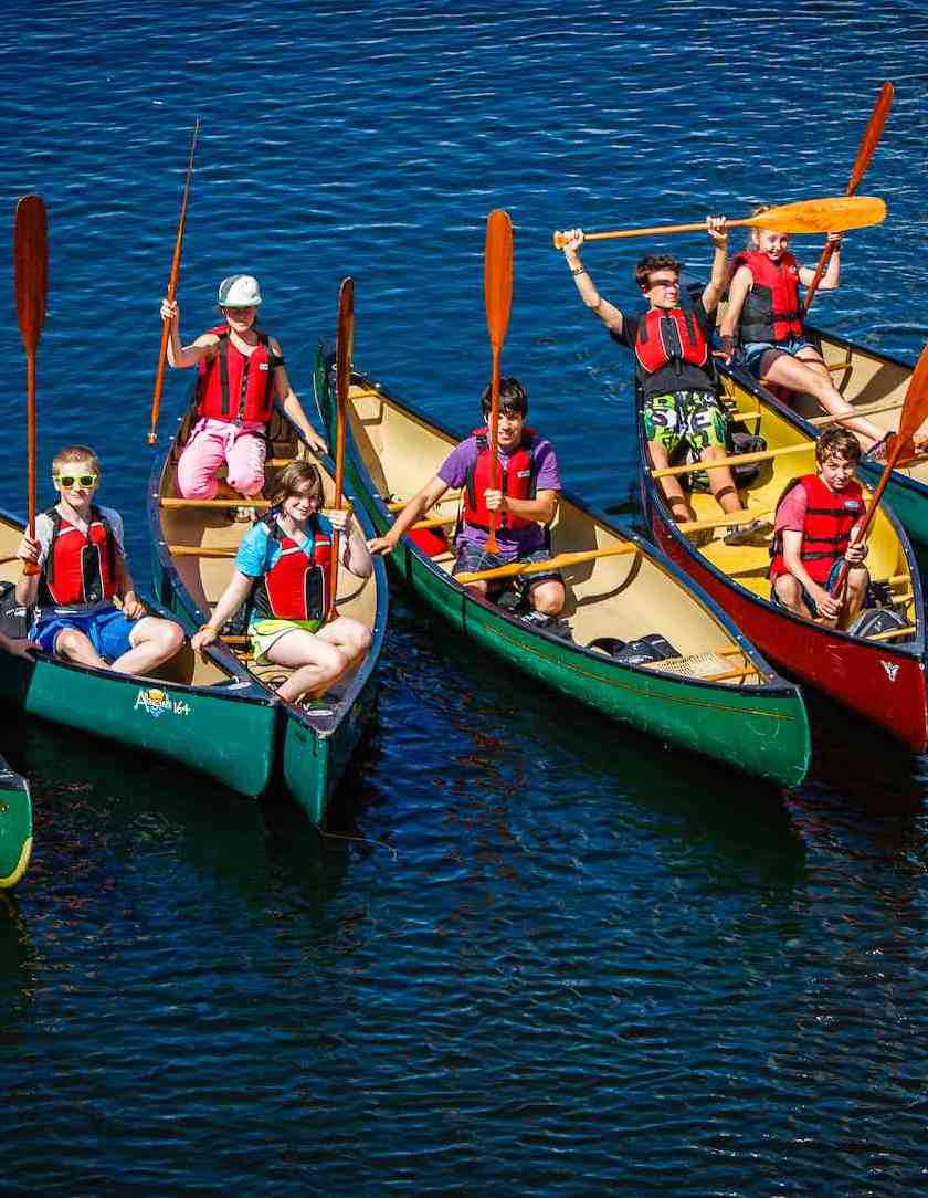 Campers and their instructors all raise their paddles to the camera in seven side by side canoes.
