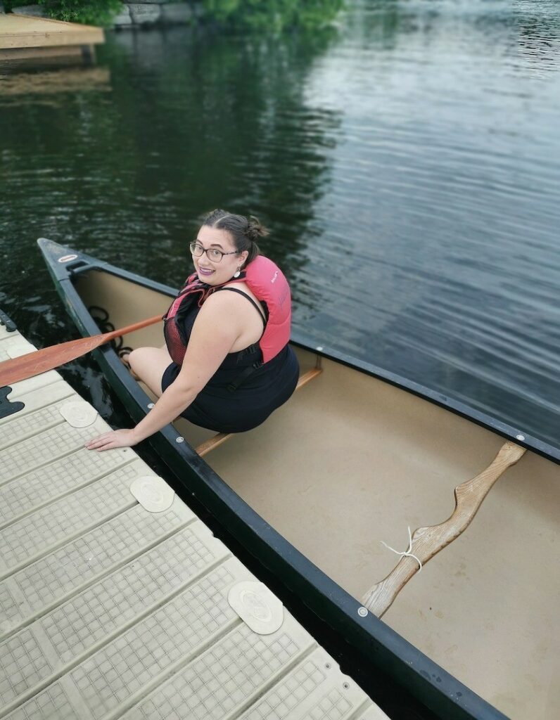 A woman wearing a personal flotation device is seated at the front of a canoe, holding a paddle and looking back over her shoulder towards the camera while bracing herself on a floating dock.
