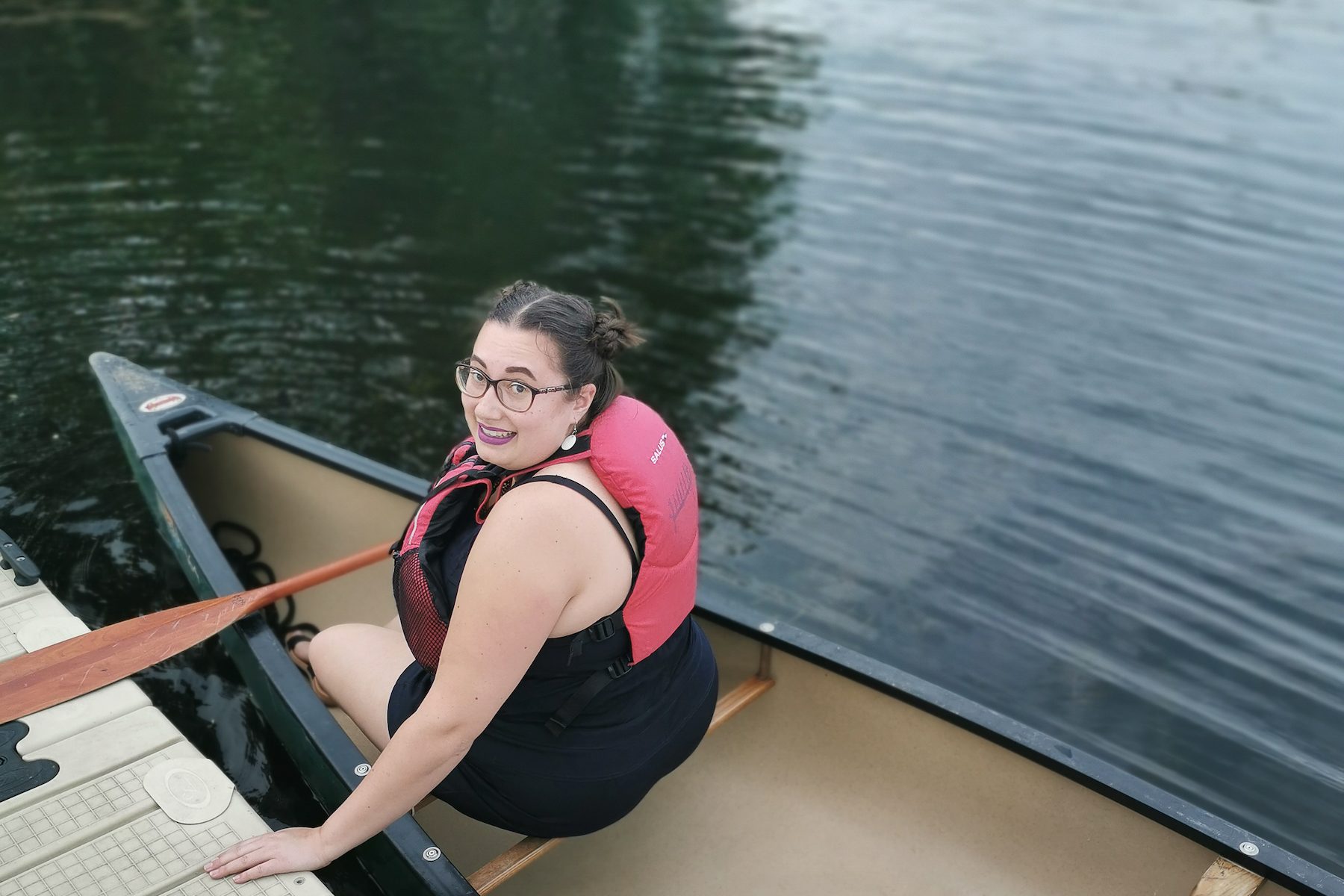 A woman wearing a personal flotation device is seated at the front of a canoe, holding a paddle and looking back over her shoulder towards the camera while bracing herself on a floating dock.