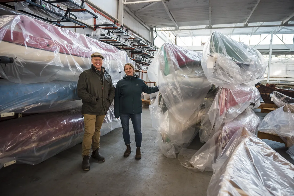 Two people stand in a storage room surrounded by canoes wrapped in plastic.