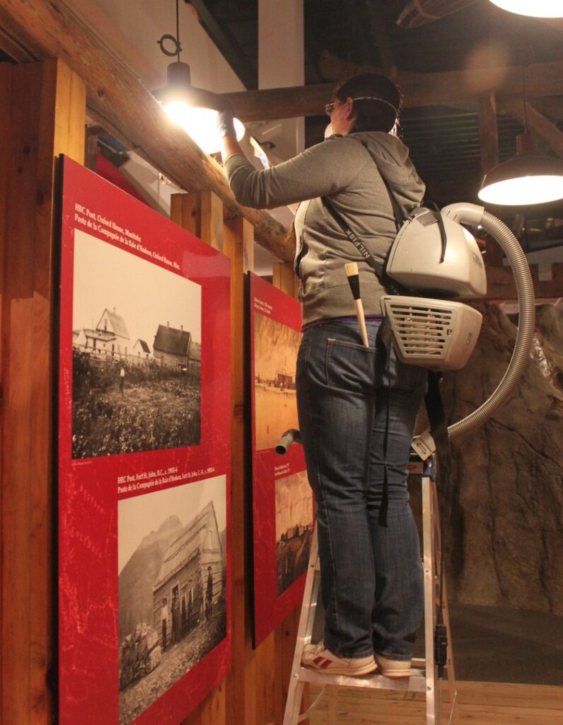 A staff member stands on a ladder with a vacuum backpack to reach high-up dust above the displays.