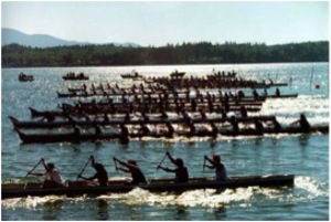 Several long canoes with over 10 paddlers each race through calm blue lake water.