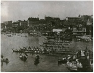 Old black and white photo of a harbour with several long canoes setting up to begin a race.