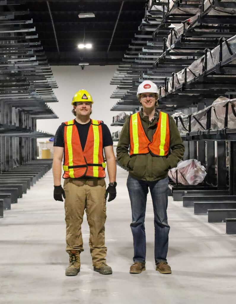 Two collections assistants wearing helmets and high visibility vests stand in an aisle of the Canadian Canoe Museum's new Collections Hall. One side of the aisle shows canoes stacked on racking. The other side is empty.