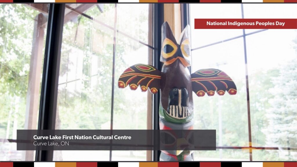 A colourful totem pole stands before large glass windows, with trees in the background, at the Curve Lake First Nation Cultural Centre in Curve Lake, Ontario. Text overlays says: National Indigenous Peoples Day.