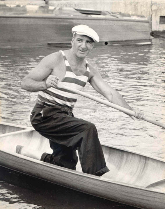 Doc Whittal wears a striped tank and white hat as he kneels in a canoe.