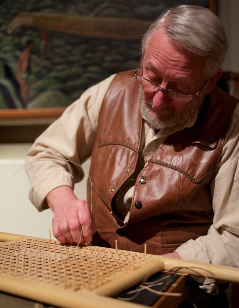 Don Duncan canes a canoe seat while sitting in the galleries of The Canadian Canoe Museum