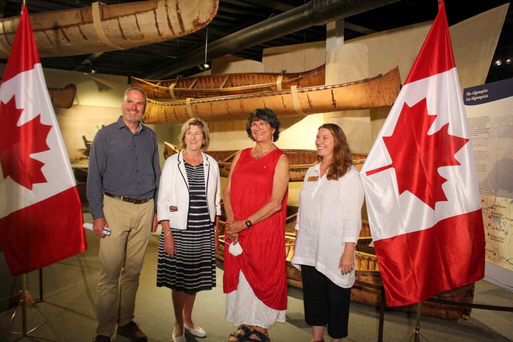 Four individuals pose for a photograph between two Canadian flags.