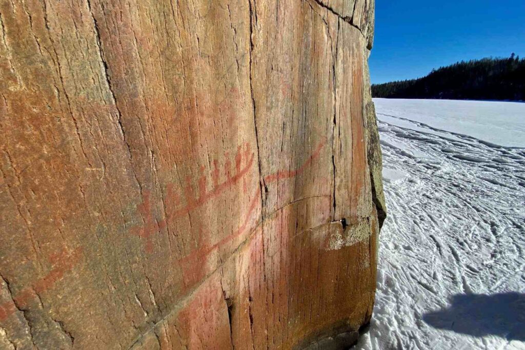 A close-up of the red mazinaawbikinigin (rock painting) that is featured in The Canadian Canoe Museum’s logo illustrating eight people in a canoe with long paddles at the bow and stern. Snow covers the ground.