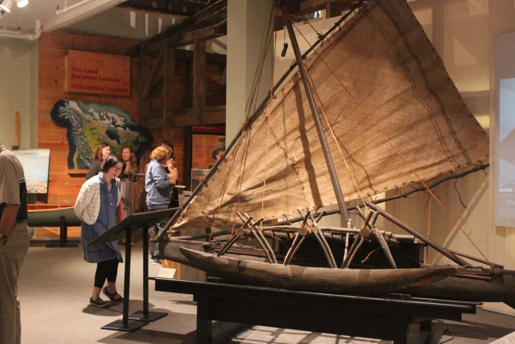 Visitors in an exhibit stand in front of an old canoe with sail