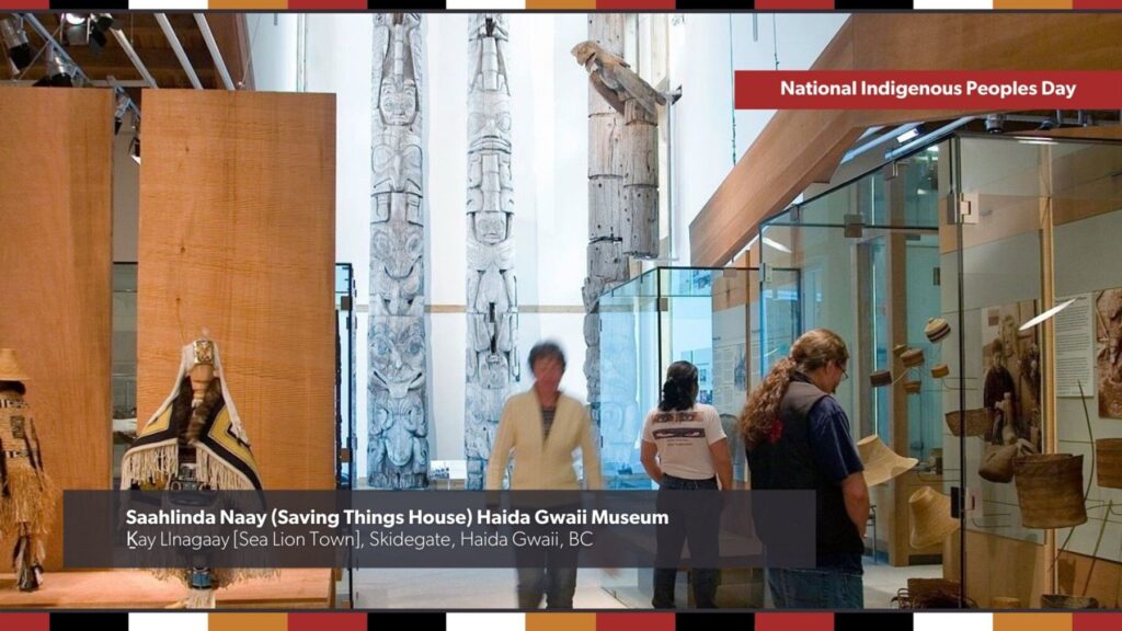 Visitors enjoying the Saahlinda Naay (Saving Things House) Haida Gwaii Museum in Kay Llnagaay [Sea Lion Town], Skidegate, Haida Gwaii, BC. In the foreground, visitors are looking at cultural objects and artifacts behind glass, while mannequins wearing regalia are off the side and large wood carvings that reach the ceiling are shown in the background. Text overlay says: National Indigenous Peoples Day.