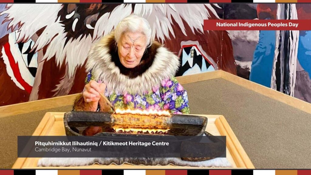 An older woman lights a qulliq, a traditional oil lamp at Pitquhirnikkut Ilihautiniq / Kitikmeot Heritage Centre in Cambridge Bay, Nunavut. Text overlay says: National Indigenous Peoples Day.