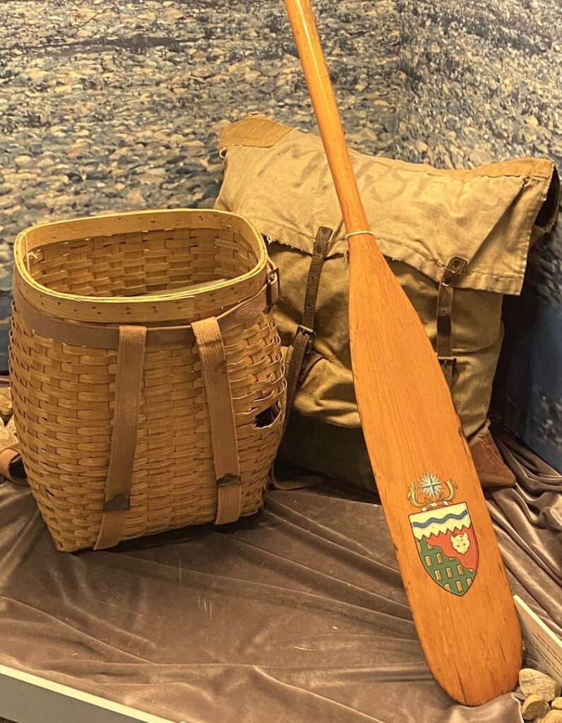 Display of weaved basket, sack, and painted wooden canoe paddle.