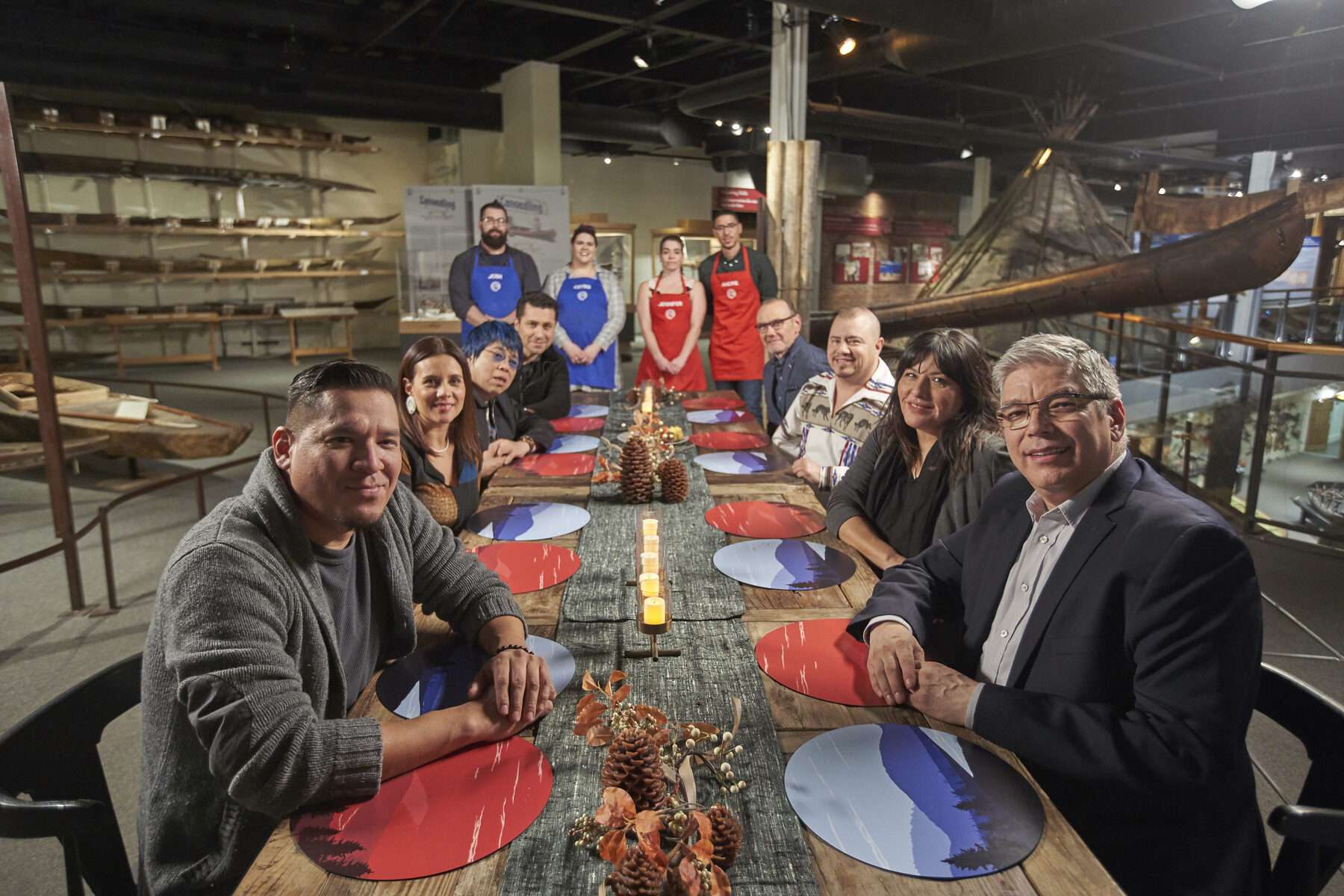 Several people sit at a harvest table with four chefs at the head of the table wearing MasterChef aprons