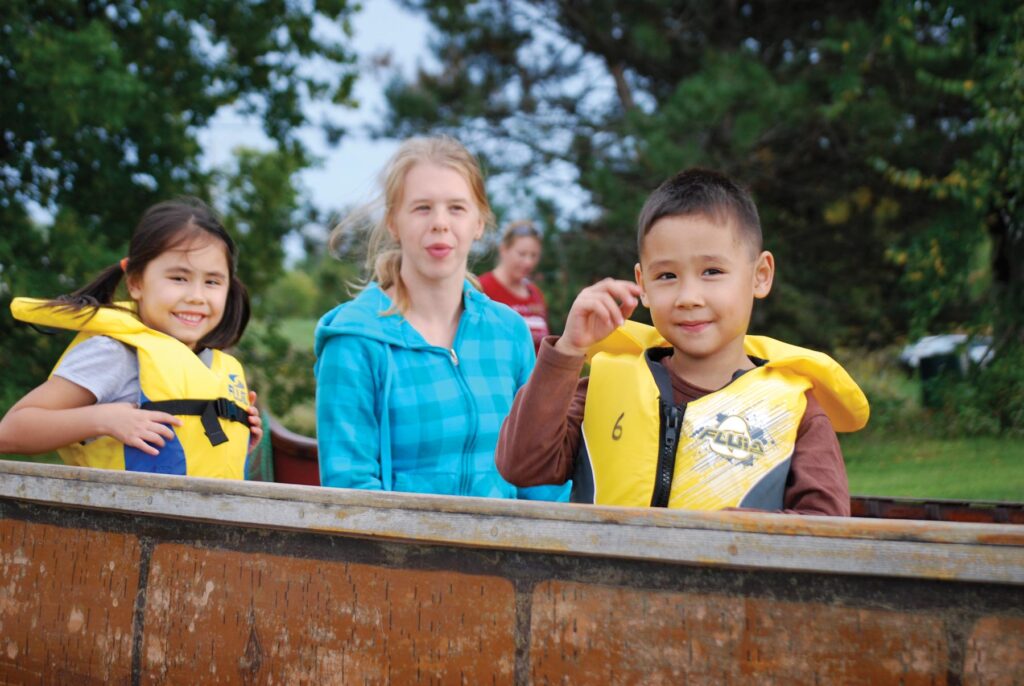 Children stand smiling beside a birch bark canoe outside.