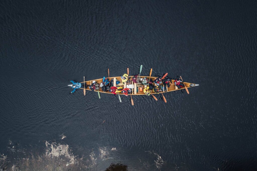 A group of paddlers in a large voyageur canoe look up to the sky at the camera.