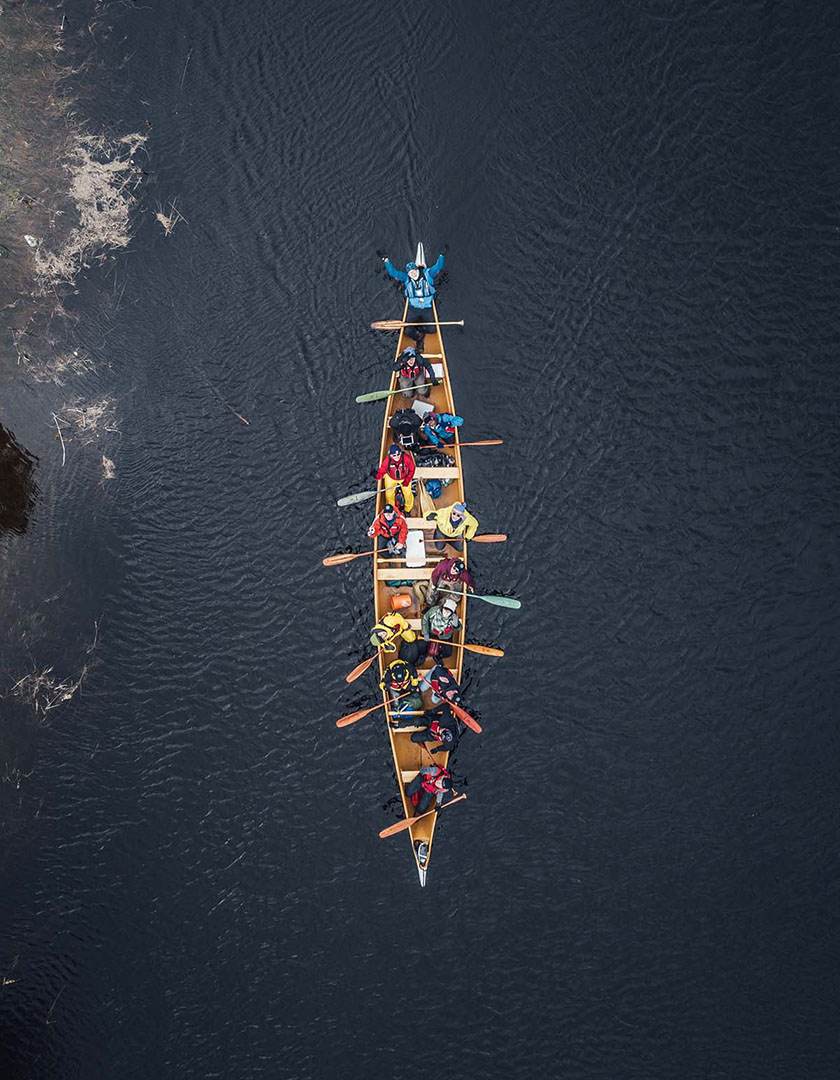 A group of paddlers in a large voyageur canoe look up to the sky at the camera.