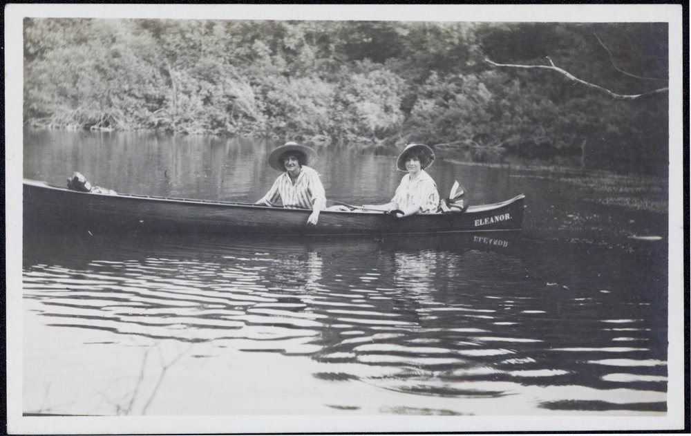 Postcard of a black and white photograph. Two women float in a long canoe with a forest in the background and calm water surrounding.