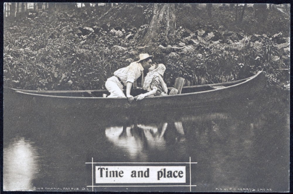 Postcard of a black and white image. A canoe floats in calm water next to a lush shore and forest while a man and woman kiss.