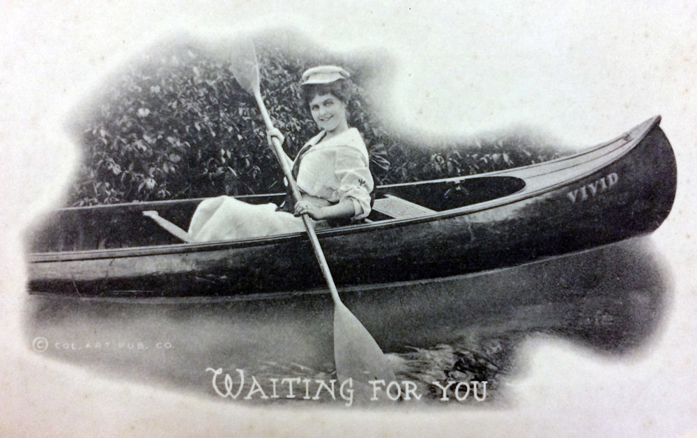 Postcard of a black and white photograph. A woman smiles while holding a kayak paddle and sits in a canoe named "Vivid".