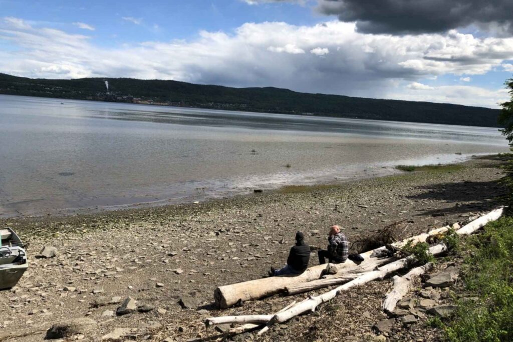 Two men sit on beached driftwood while looking out at the water