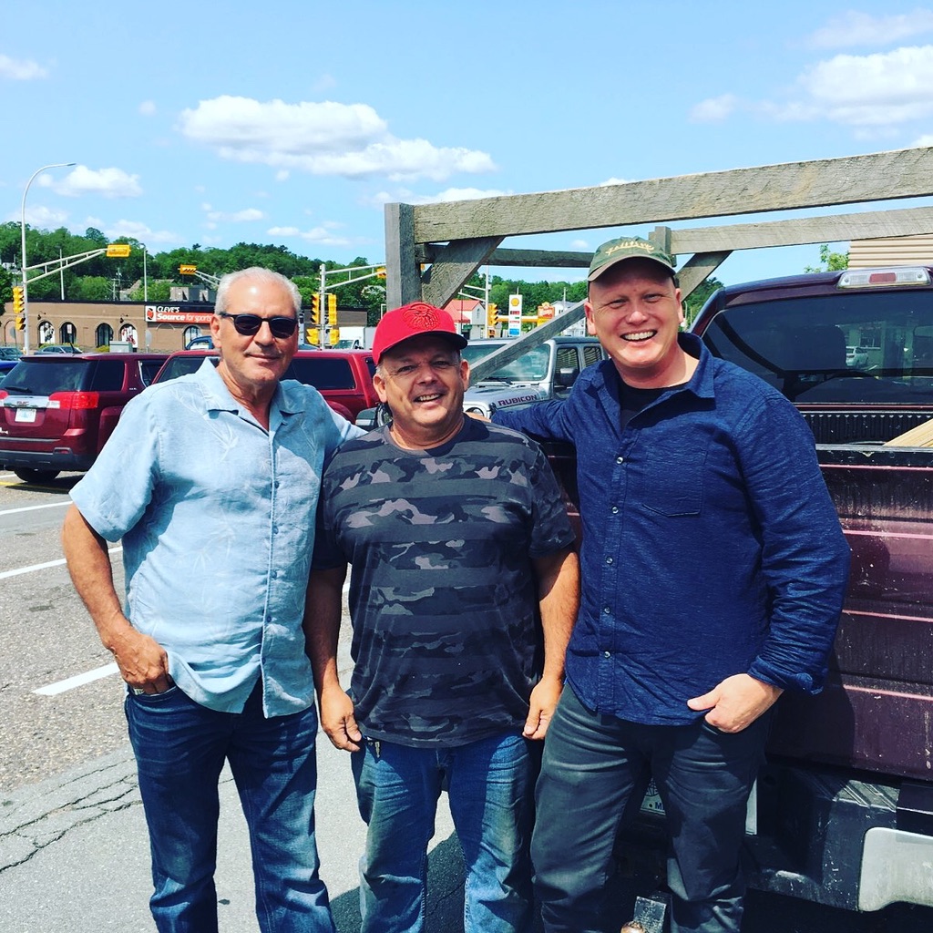 Three men smile for a picture while leaning against a truck in a parking lot