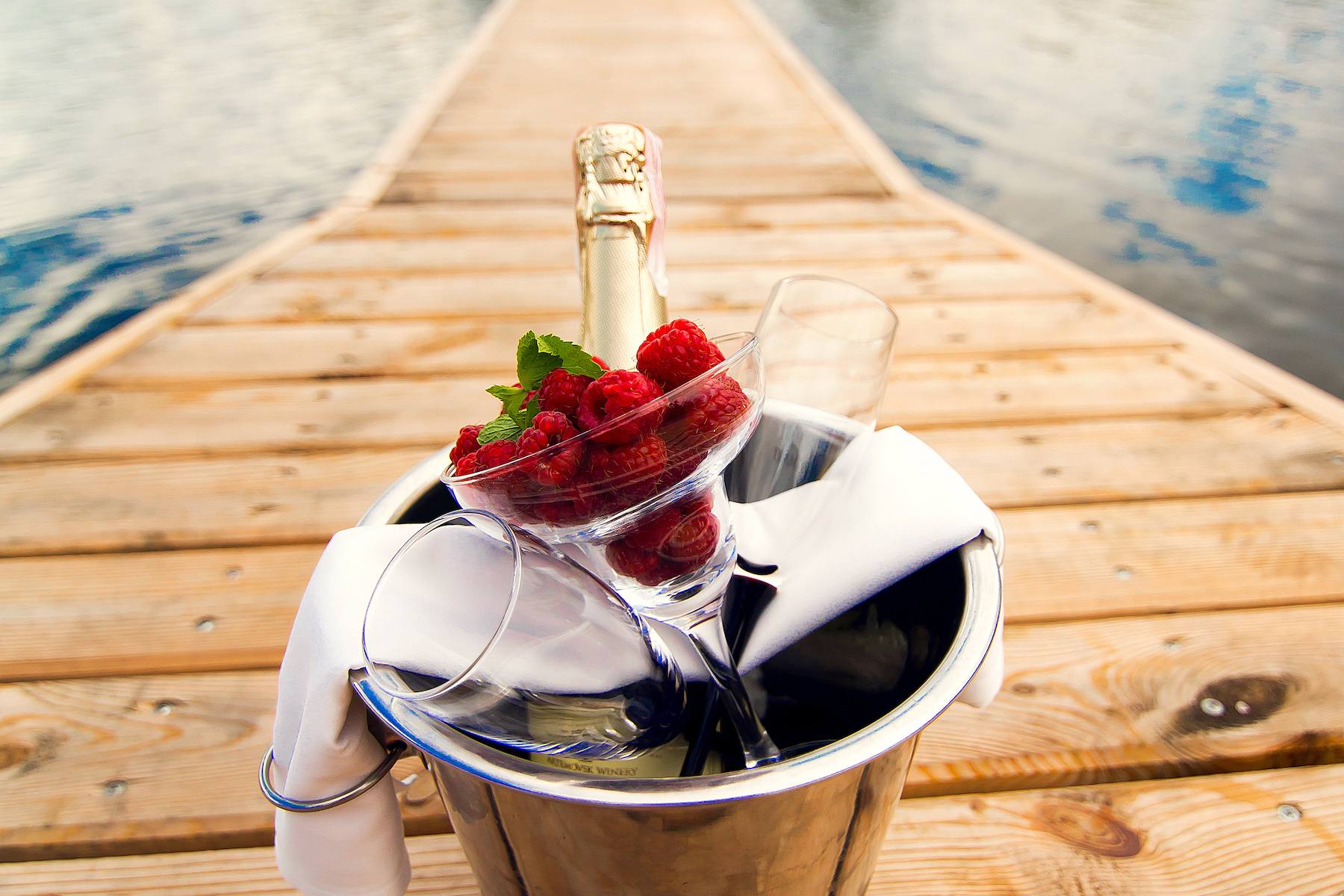 An ice bucket with two champagne fluets, a bottle of champagne, white cloth, and a martini glass filled with raspberries sits on a wooden dock on a lake. Water and trees are in the background.