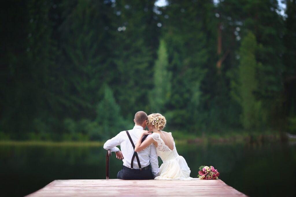 A bridge and a groom sit at the end of a wooden dock on a lake with their backs to the camera. The bride leans on the groom. Her bouquet sits on the dock beside her. In the background are trees.
