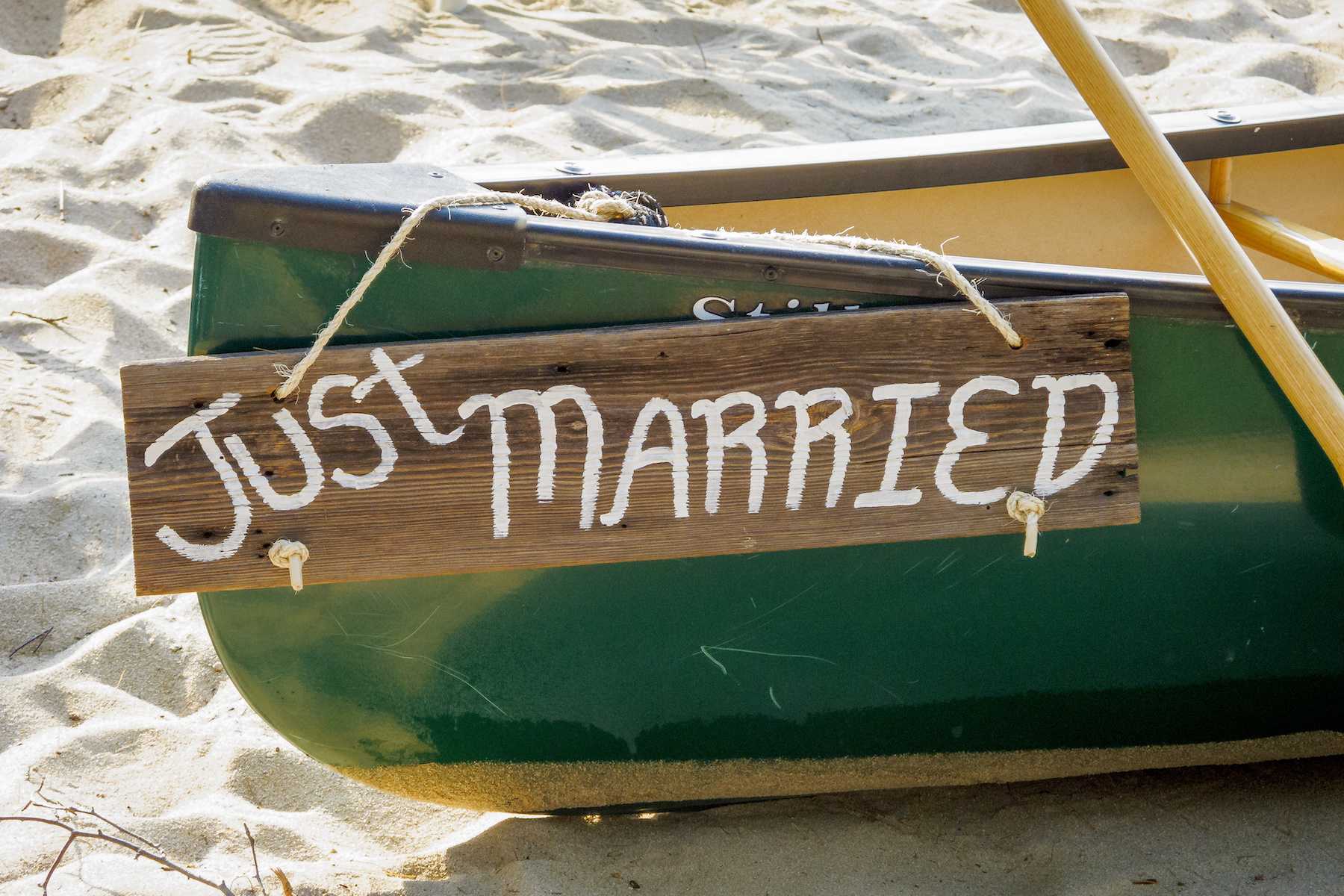 A green canoe rests on a sandy shoreline. A wooden plaque is tied to the stern with rope. It says Just Married.