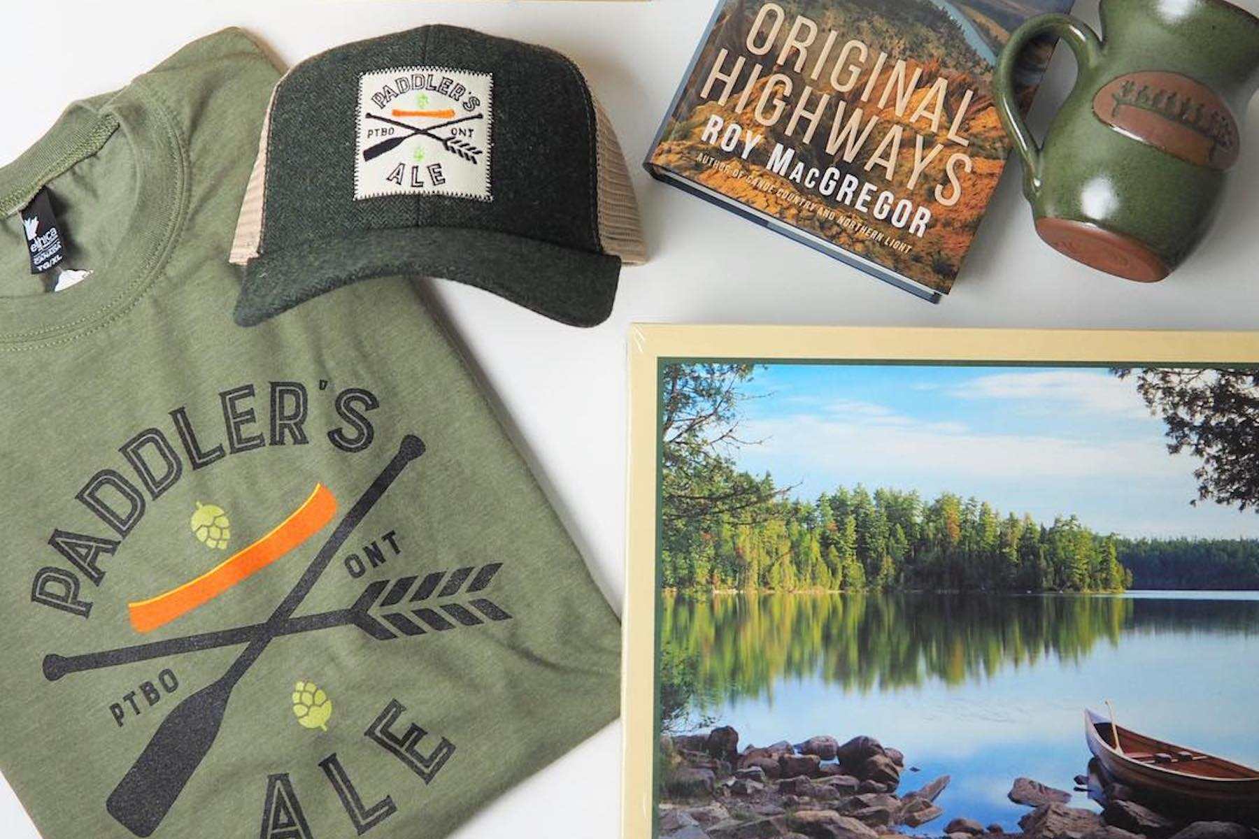 A green Paddler's Ale t-shirt and baseball hat, a wooden canoe paddle rack, Original Highways by Roy McGregor, a ceramic mug with The Canadian Canoe Museum's logo, and a puzzle of a canoe on a lake.
