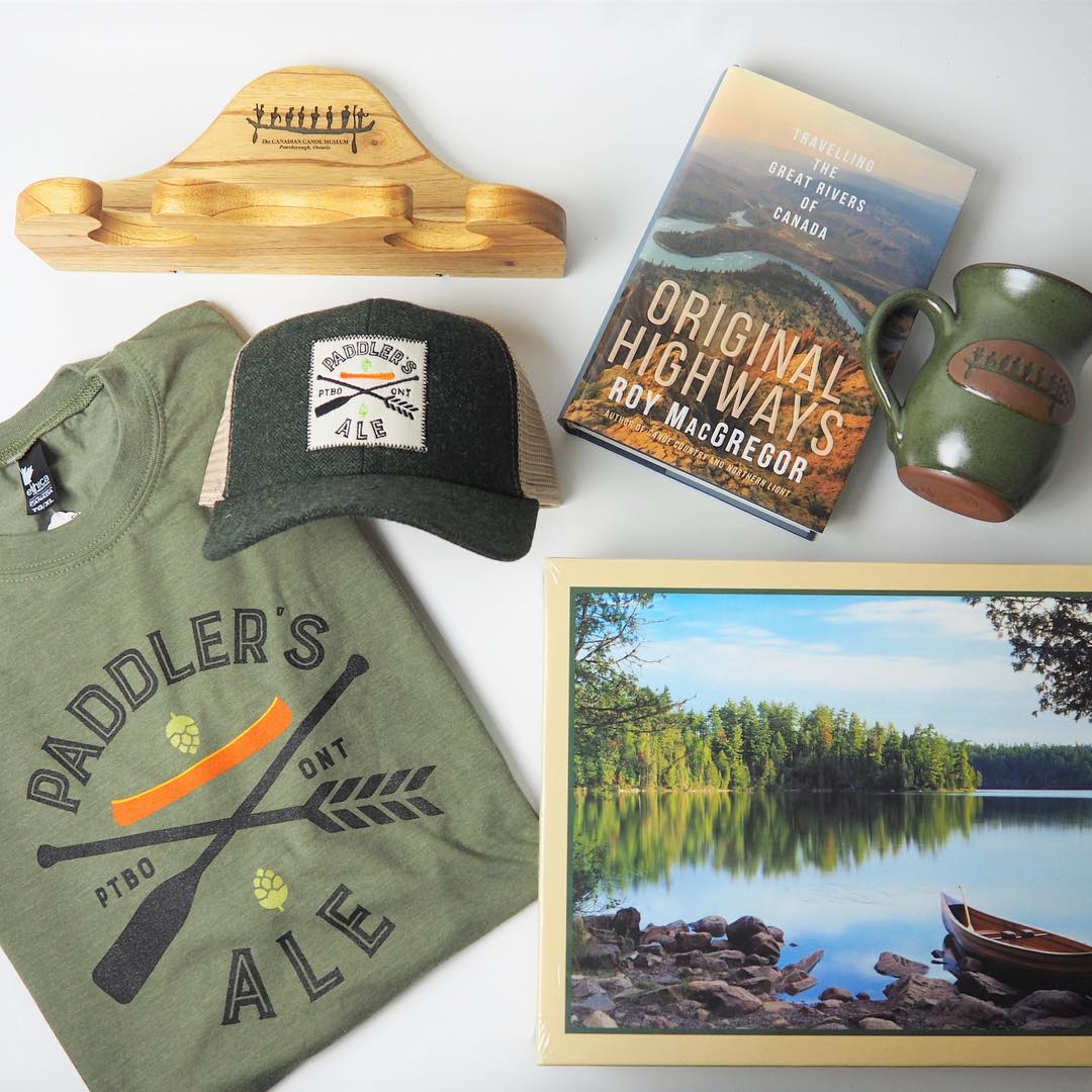 A green Paddler's Ale t-shirt and baseball hat, a wooden canoe paddle rack, Original Highways by Roy McGregor, a ceramic mug with The Canadian Canoe Museum's logo, and a puzzle of a canoe on a lake.