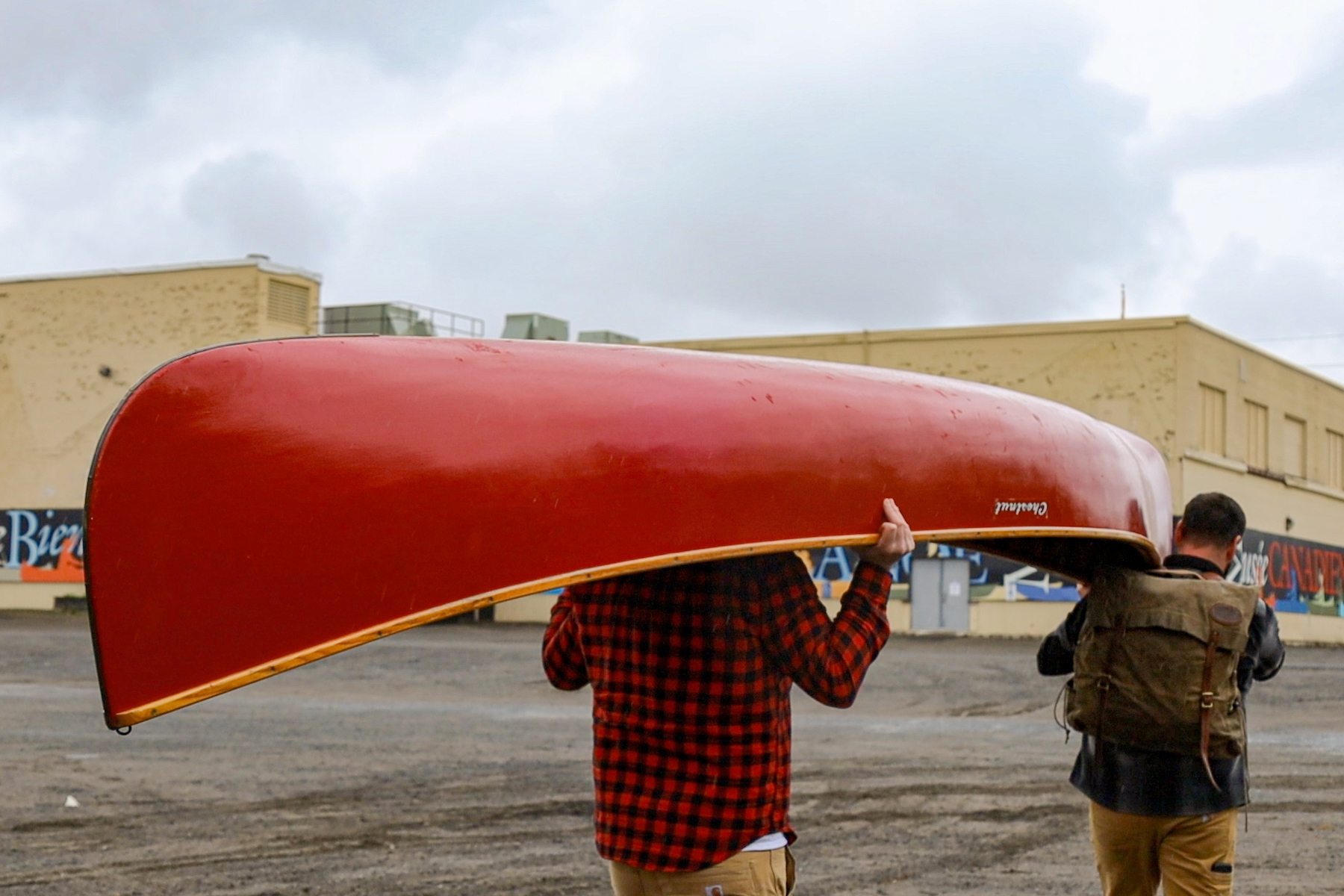 Two men in plaid portage or carry a red Chestnut canoe across The Canadian Canoe Museum's parking lot at 910 Monaghan Road.