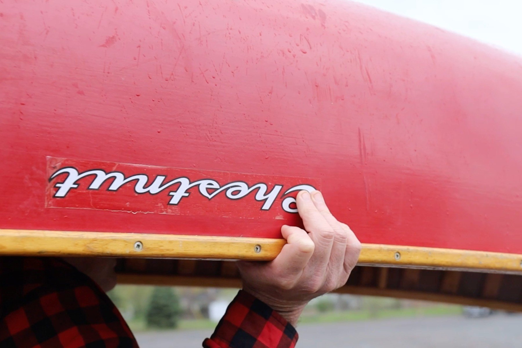 A man in red plaid grips the gunwales of a red Chestnut canoe while portaging it.