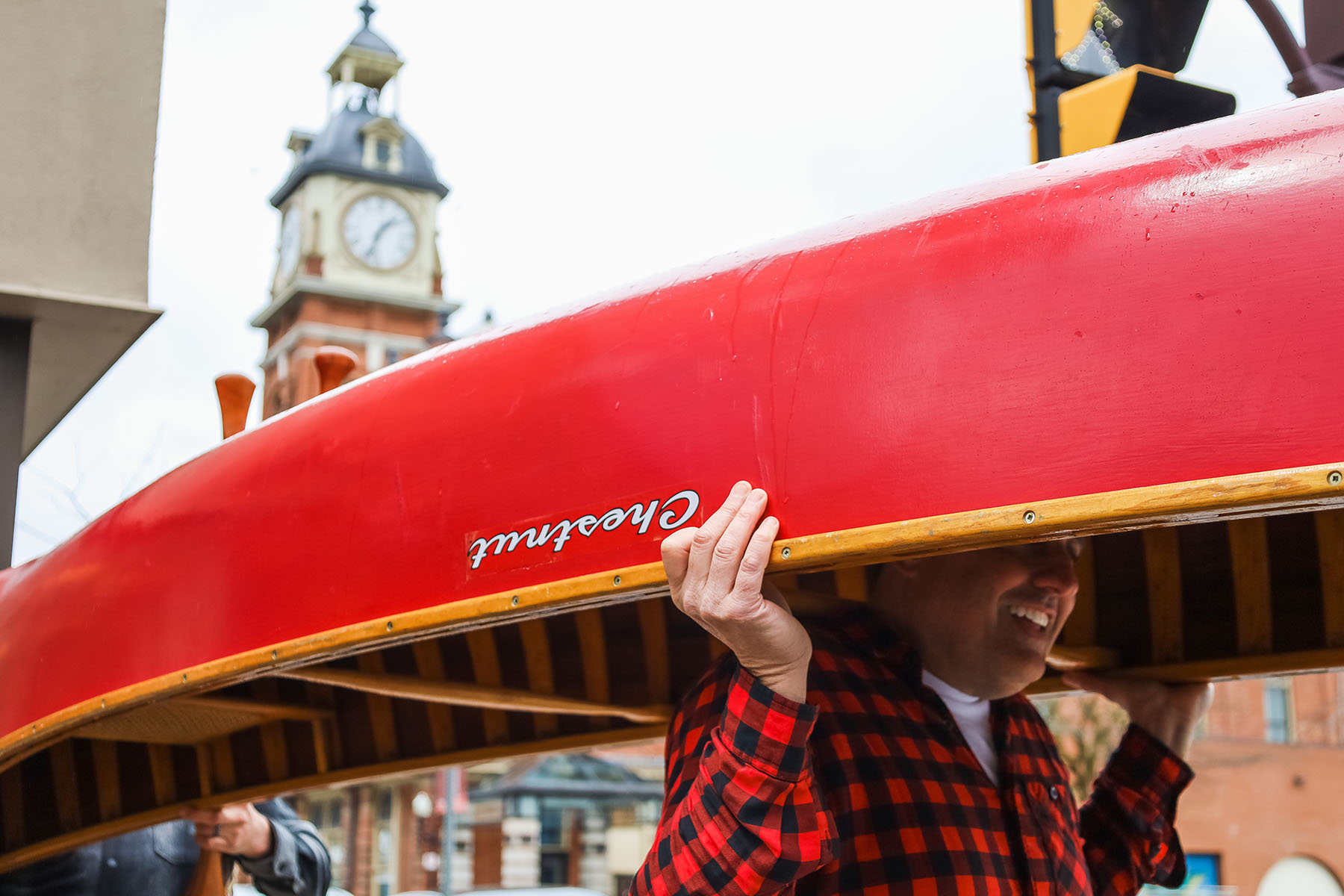 A man in red and black plaid portages or carries a red Chestnut canoe in downtown Peterborough with the Market Hall clock tower in the background.