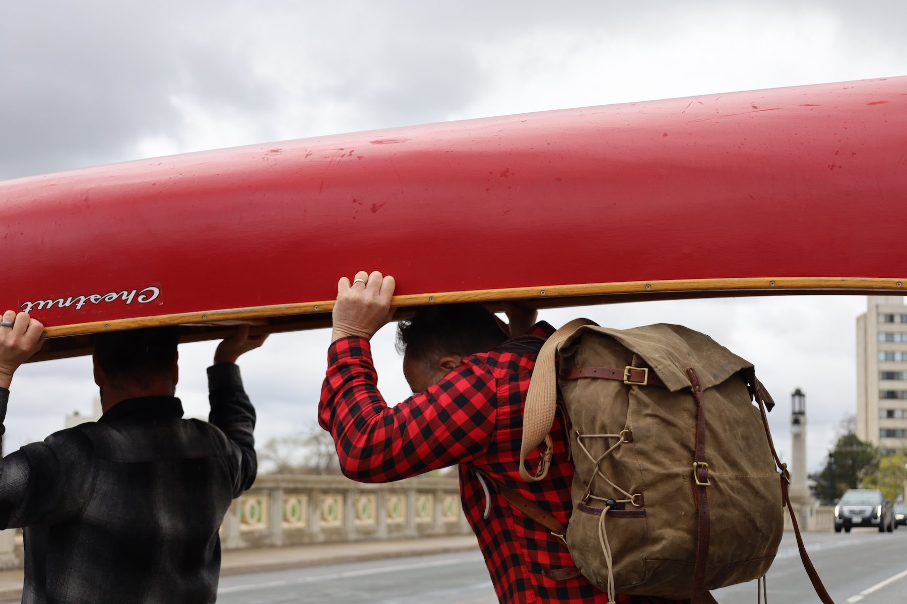 Two men in plaid portage or carry a red Chestnut canoe across the Hunter Street Bridge in East City, Peterborough.