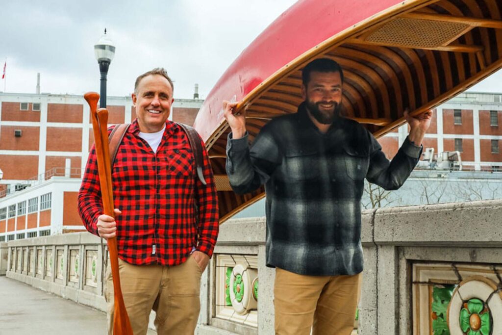 Neil Morton and Mike Judson stand smiling on the East City bridge in Peterborough, Ontario, both wearing plaid. Neil is wearing a pack and carrying two wooden paddles. Neil is portaging a red Chestnut wooden canoe.