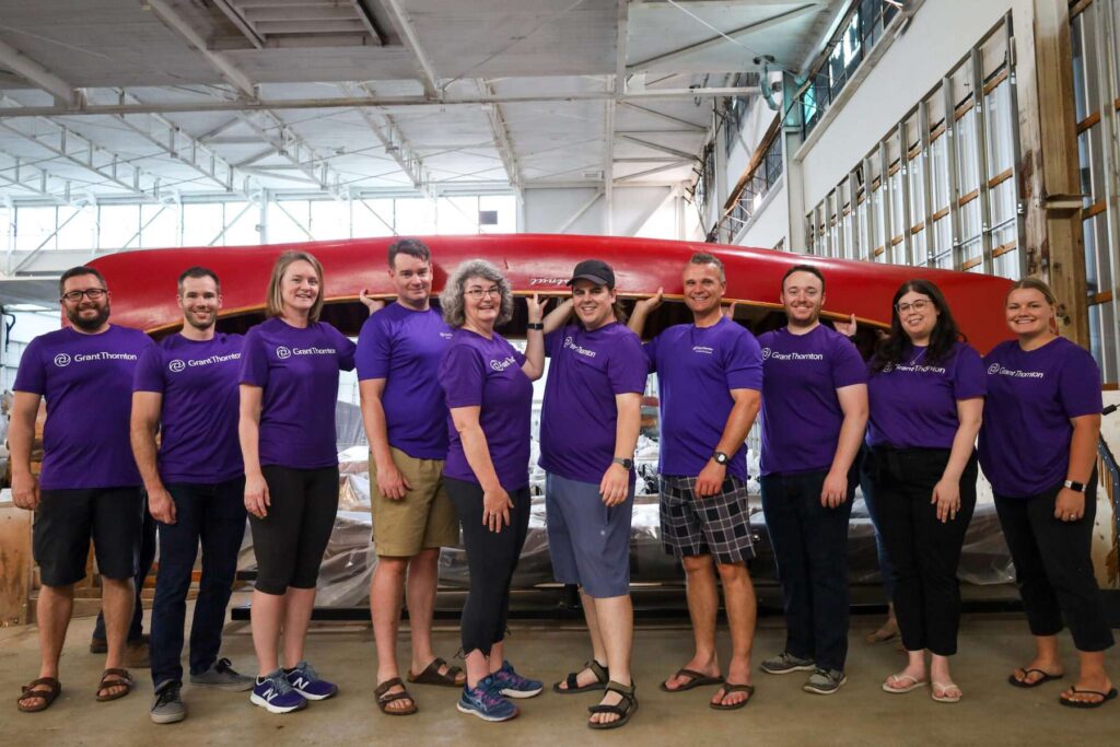 Ten Grant Thorton staff members smile at the camera while  lifting up a red Chestnut wooden canoe in the Canadian Canoe Museum's old Collection Centre at 910 Monaghan Road. They are all wearing purple shirts with the Grant Thorton logo in white.
